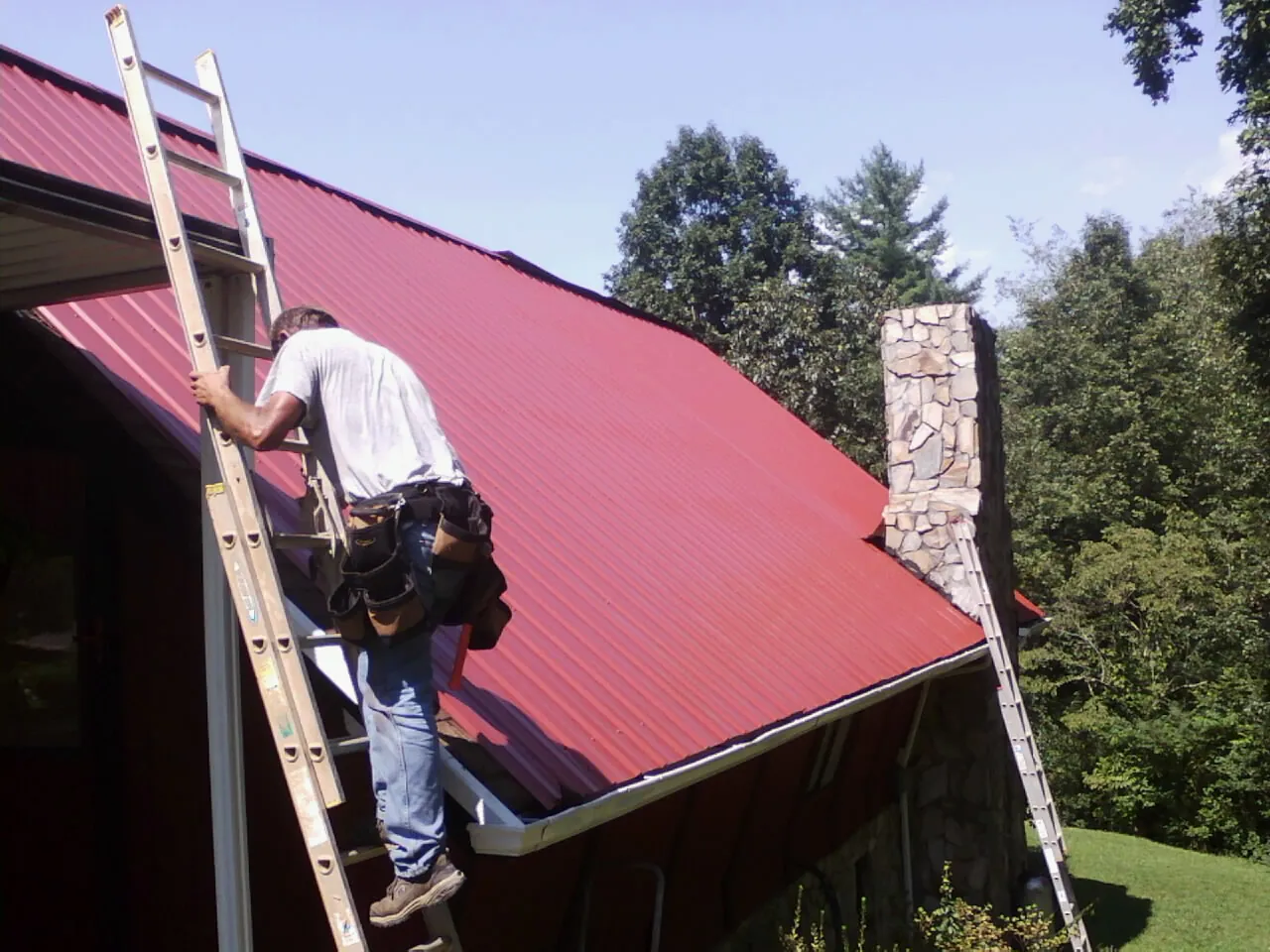Roofer on a ladder beside a red metal roof and stone chimney — Soffit & Fascia Repair by Roofing Brothers in Wapwallopen
